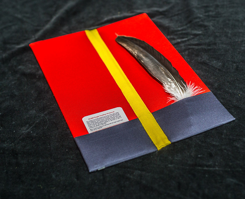 An eagle feather is sitting in a red and blue folder; the folder has a yellow ribbon around it.