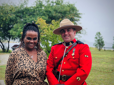 A male RCMP officer in red serge is standing beside a woman outdoors; both people are smiling at the camera.