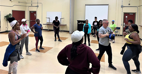 A plain-clothes RCMP officer and a woman are demonstrating a self defense move while eleven other women look on.