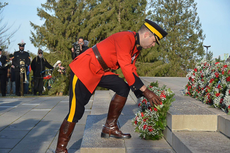 A male RCMP member in red serge is leaning over to place a wreath at the foot of a monument. Other people and wreaths are also pictured.