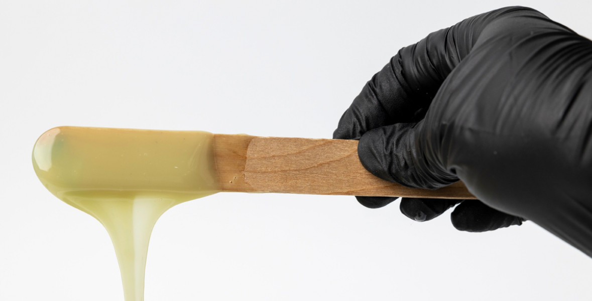 A black-gloved hand holds a stick dripping with light-yellow body wax.