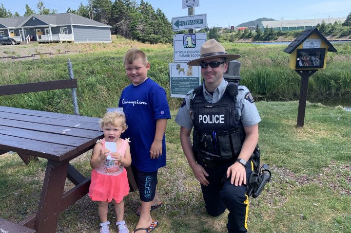 A uniformed RCMP officer is kneeling next to a boy and girl beside a picnic table. The children are both holding prize tickets.