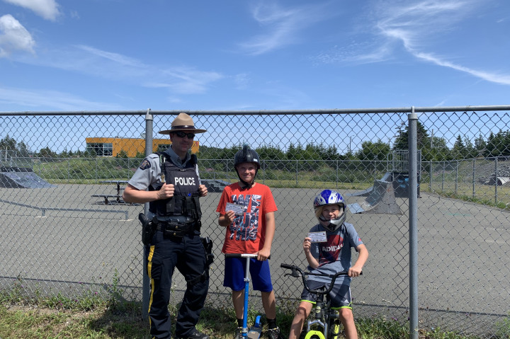 A uniformed RCMP officer and two boys on bicycles in front of a chainlink fence. All are smiling and the boys are holding prize tickets.