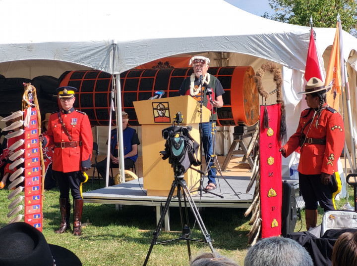 Master carver, Stanley Hunt, standing at a podium speaking to a crowd in front of the 18-foot-tall carving