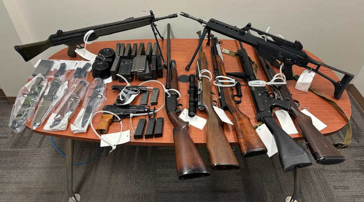 Numerous seized rifles, handguns, magazines, bayonets and a shotgun are displayed on a large table.