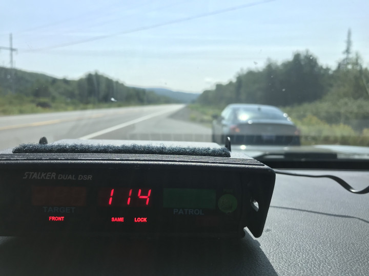 A radar device mounted on the dash of a police vehicle displays a speed of 114 km/h. A dark coloured car is stopped in front of the police vehicle near a roadway.