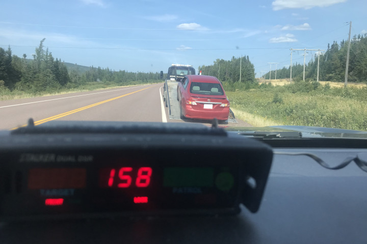 A radar device mounted on the dash of a police vehicle displays a speed of 158 km/h. A red car is stopped in front of the police vehicle near a roadway.