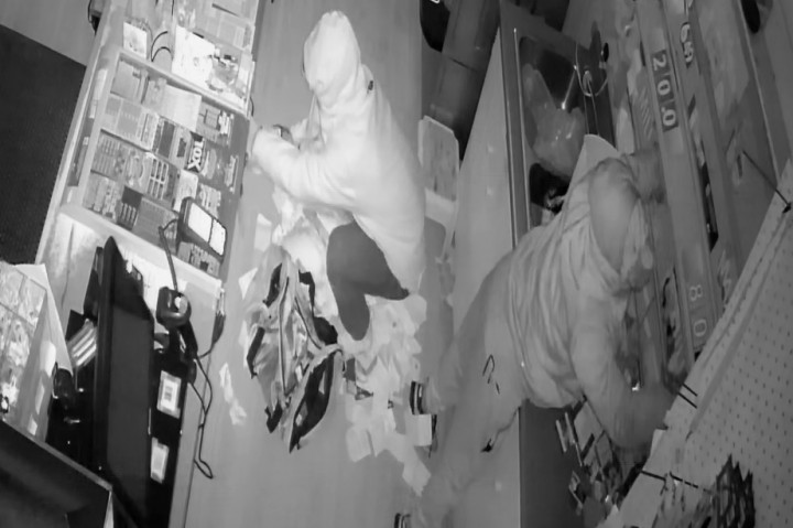 A still image taken from video surveillance footage, which shows two unknown suspects behind the cash register. One suspect is seen standing on his toes and reaching up on a shelf, while the second crouches behind the counter.