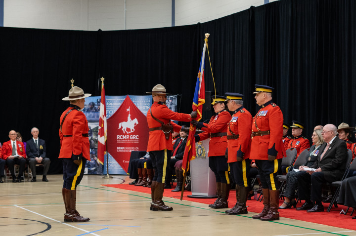 RCMP officers dressed in red serge are inside a gymnasium with people seated around them. Two officers are passing a flag.