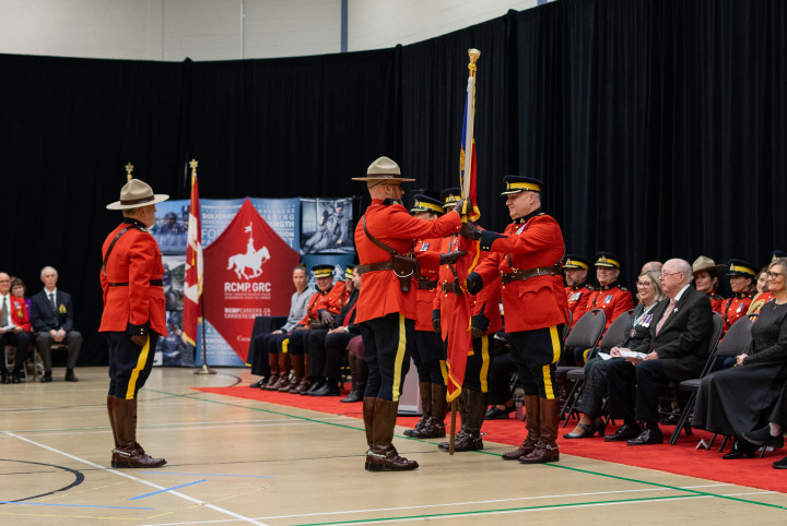 RCMP officers dressed in red serge are inside a gymnasium with people seated around them. Two officers are passing a flag.